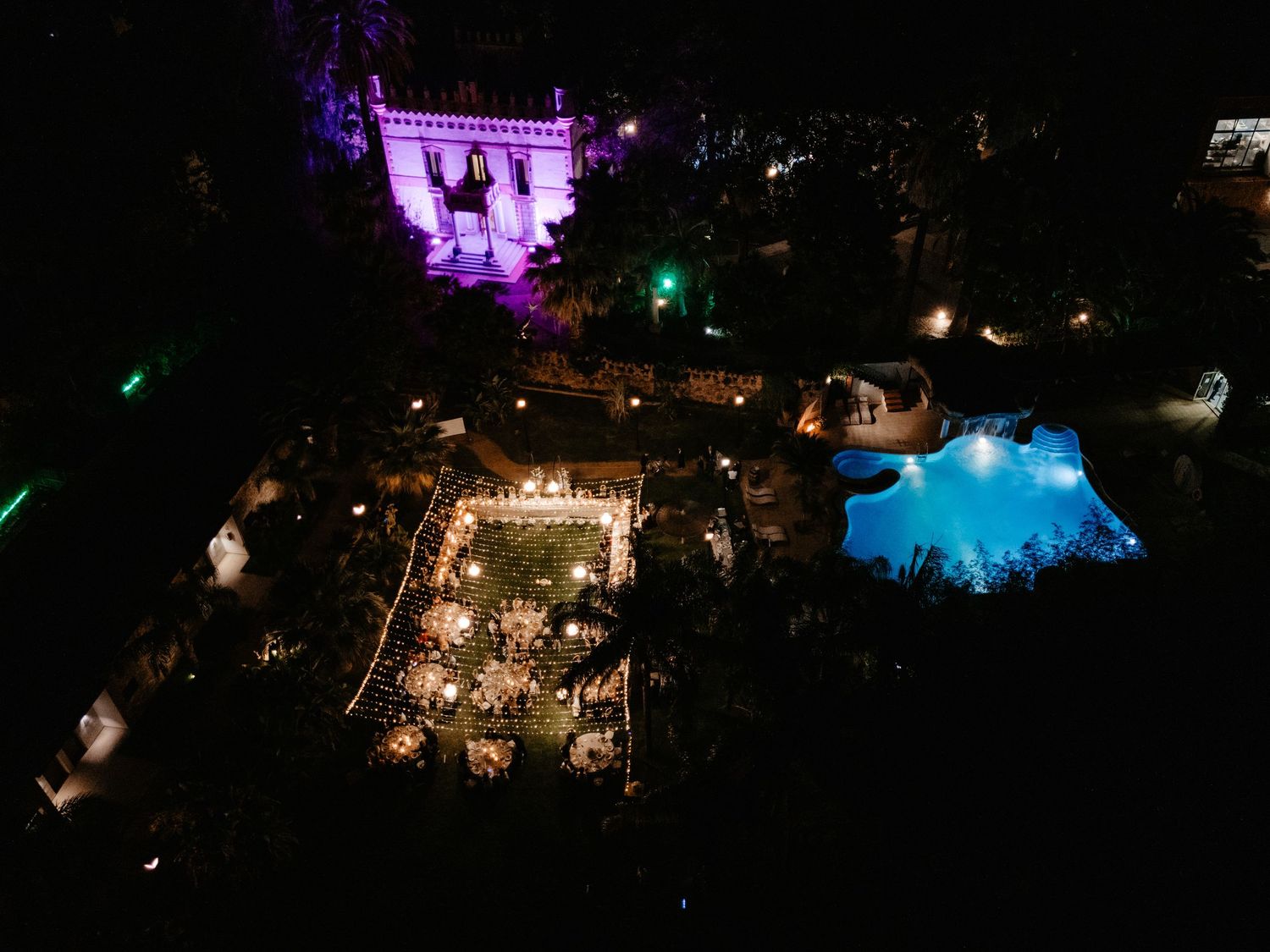 Aerial nighttime view of an illuminated outdoor venue with a glowing blue pool and purple-lit building. Wedding in Villa Retiro, Spain.