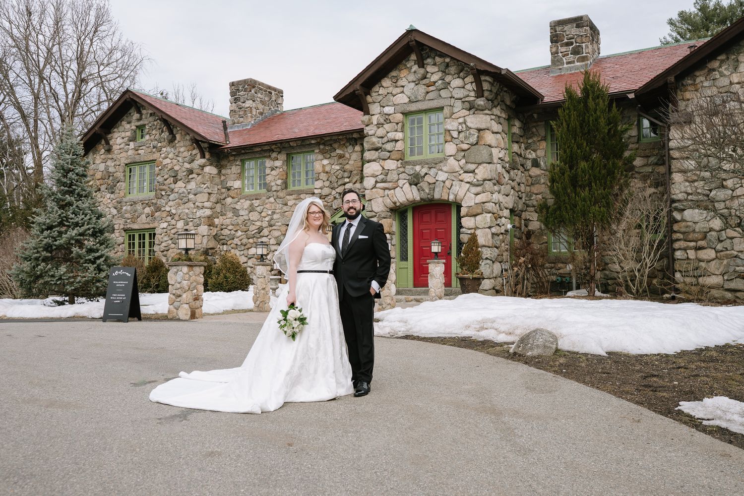 Bride and groom pose in front of a rustic stone mansion with red door and snowy grounds on their wedding day.