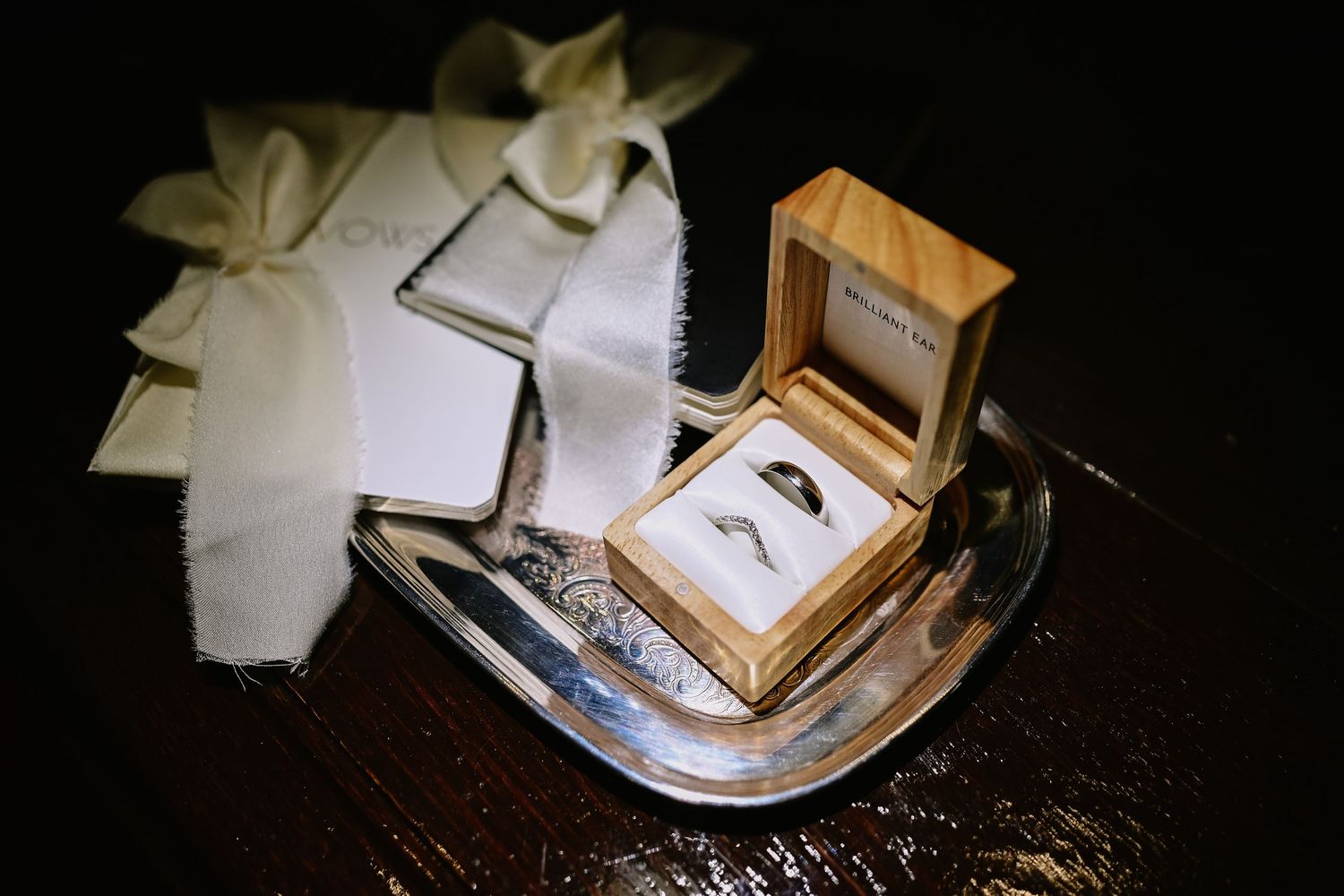 Wedding ring box with white ribbon sits on dark surface in dramatic lighting.