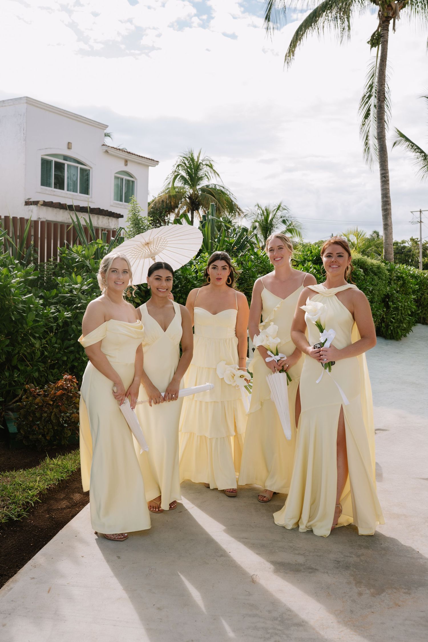 Bridesmaids in cream-colored dresses pose together on a palm-lined pathway.