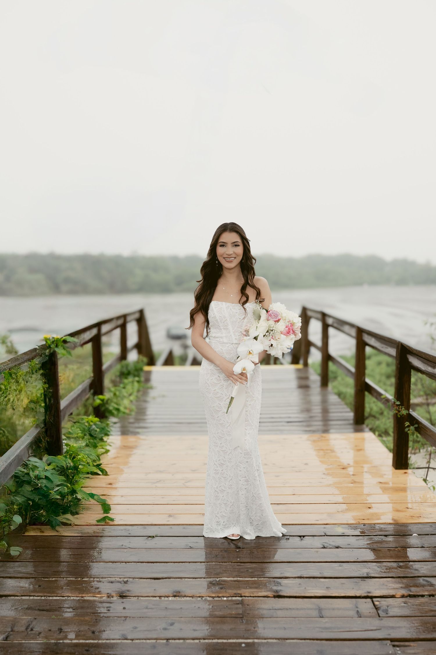 A white lace wedding dress is photographed on a wooden pier overlooking a misty lake on a rainy day.