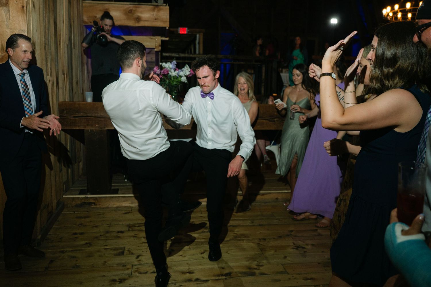 Wedding reception guests dancing and celebrating on a rustic wooden dance floor in a dimly lit barn venue.