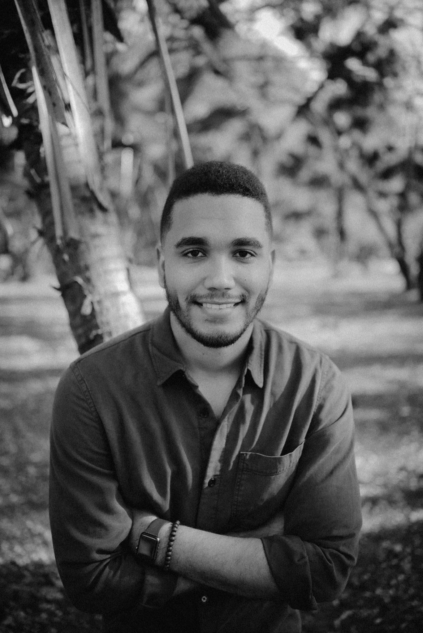 Black and white outdoor portrait of a person in casual attire smiling in a natural setting.
