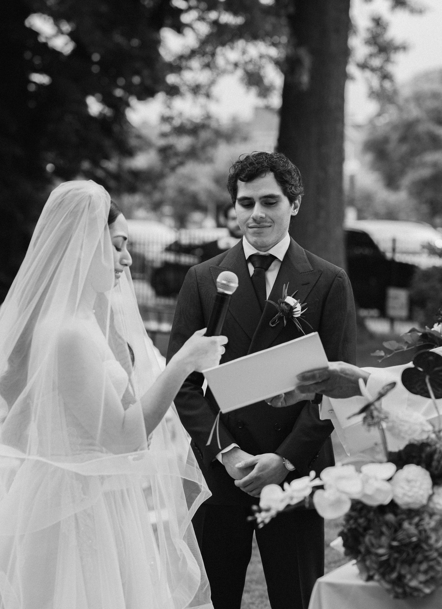 Black and white photo of a bride reading vows into a microphone while the groom smiles during an outdoor wedding ceremony.