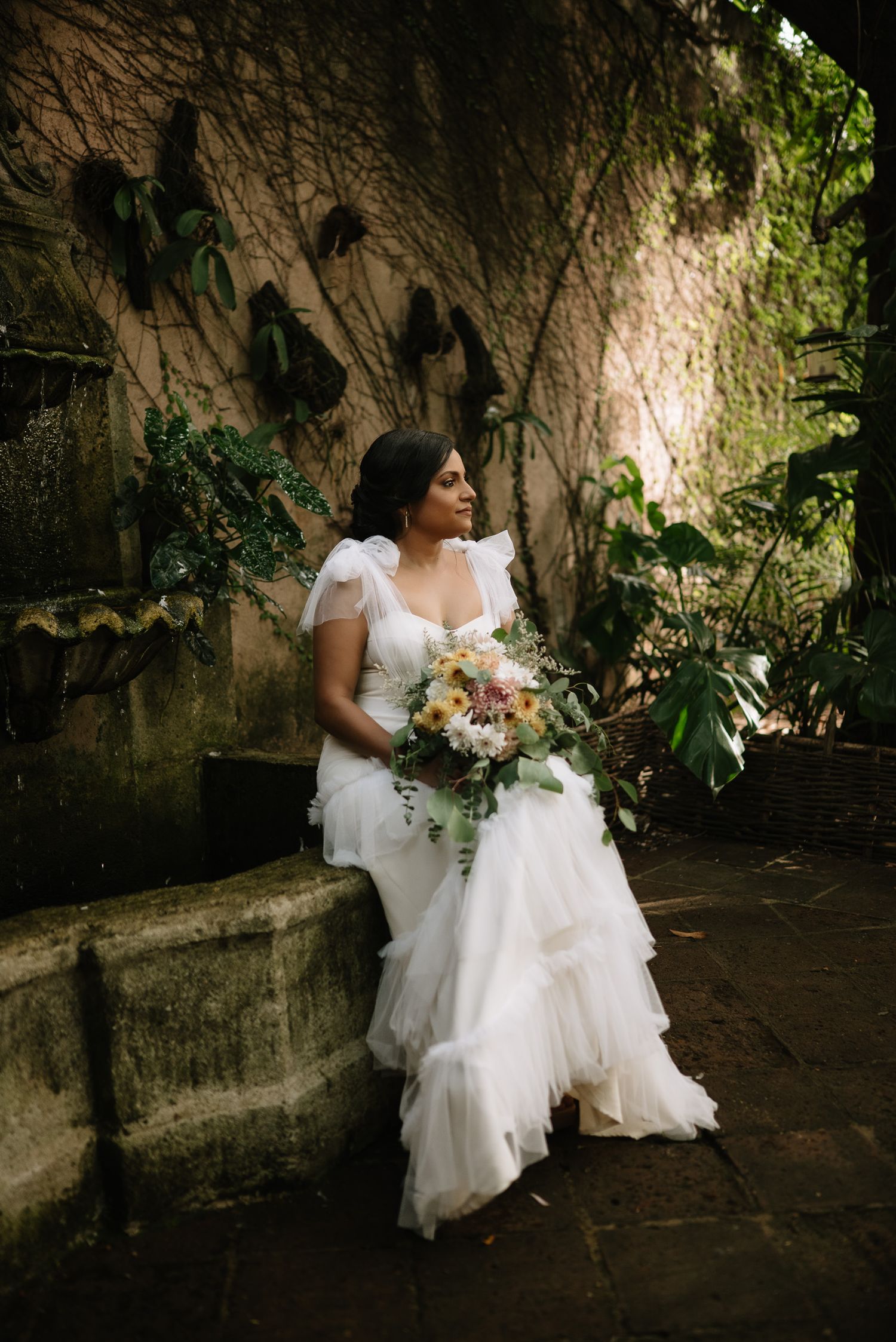 Bride in flowing white dress sits with bouquet against vintage stone wall with climbing vines.