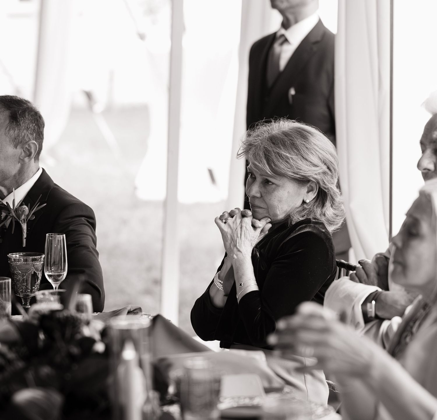 Black and white photo of a woman in dark clothing sitting at a formal dinner table, hands clasped, deep in thought.