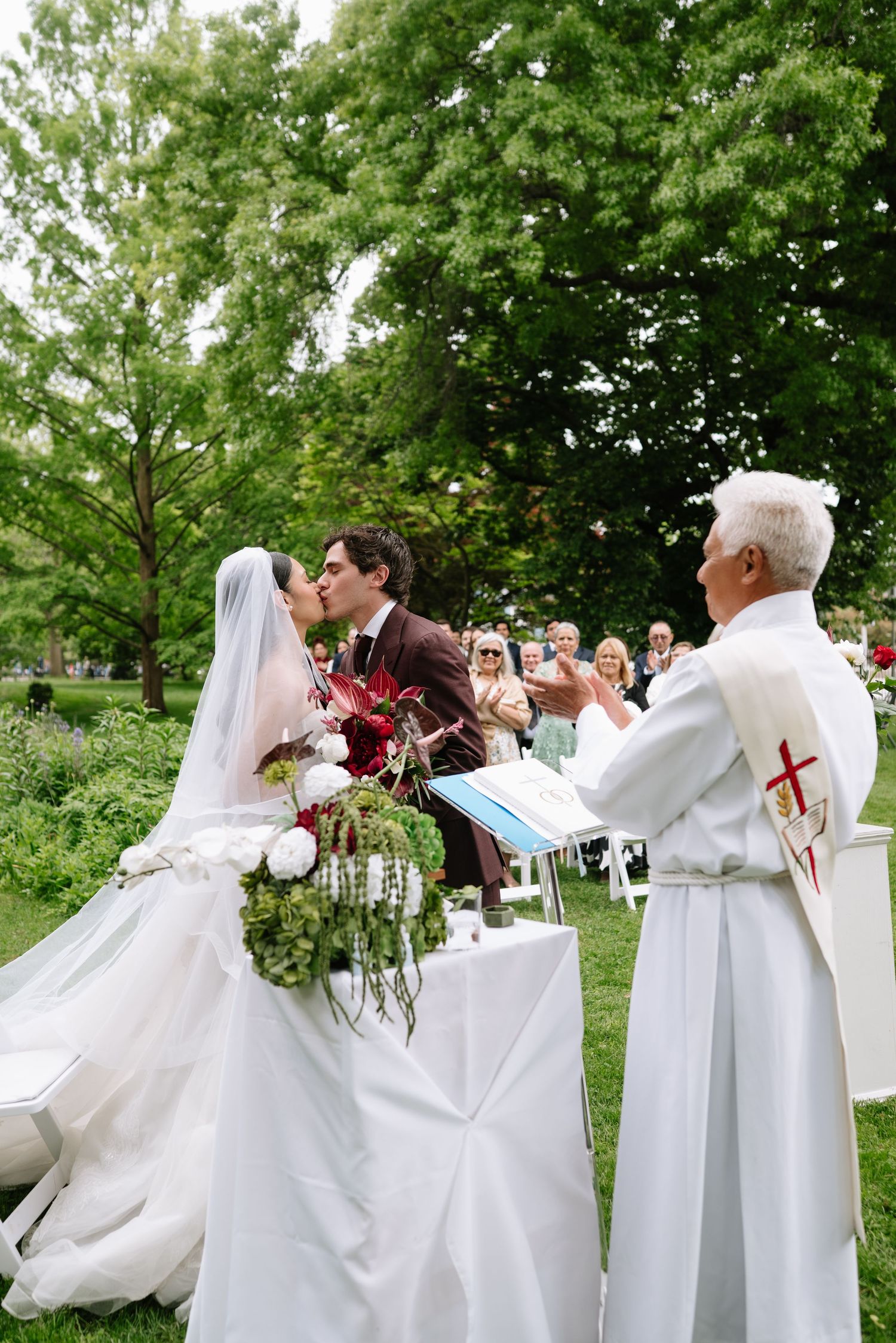 Bride and groom share first kiss at outdoor wedding ceremony while officiant applauds beside them.
