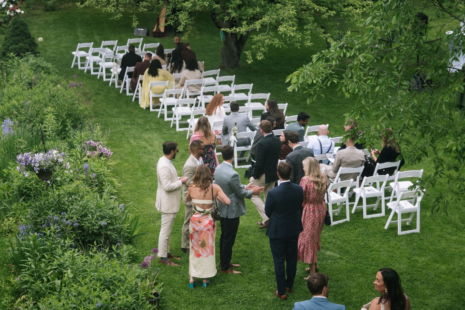 Guests gather on a lush green lawn for an outdoor wedding ceremony with white chairs arranged in rows.
