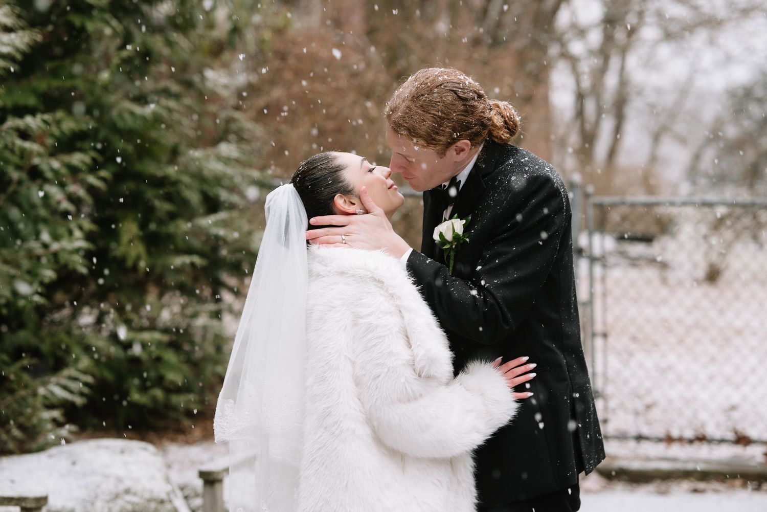 Bride and groom sharing a kiss in falling snow, bride wearing white fur wrap and veil, groom in black suit.