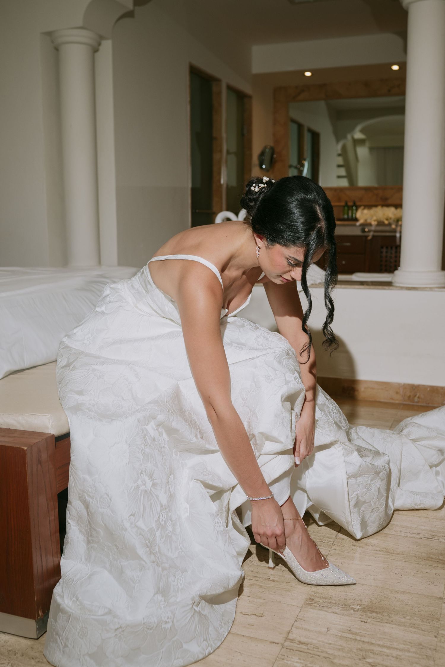 A bride in a white wedding gown adjusts her shoe while sitting on a bench.