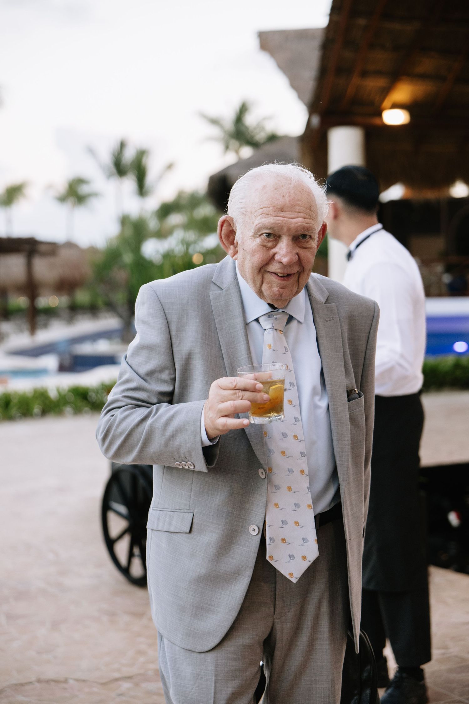 An elderly gentleman in a light gray suit and patterned tie holds a cocktail at an outdoor event.