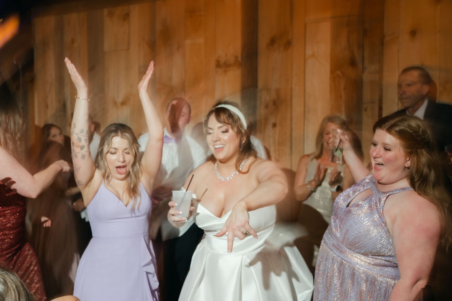 Wedding party dancing and celebrating in a rustic wooden barn venue with festive lighting.