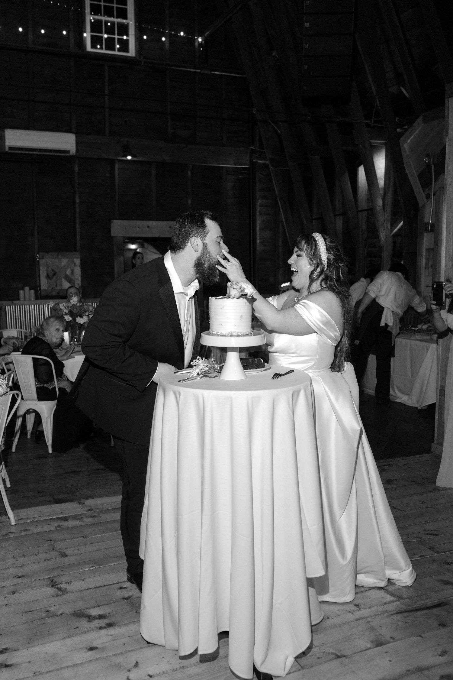 Newlyweds cut their wedding cake at a reception with white tablecloths in a rustic venue.