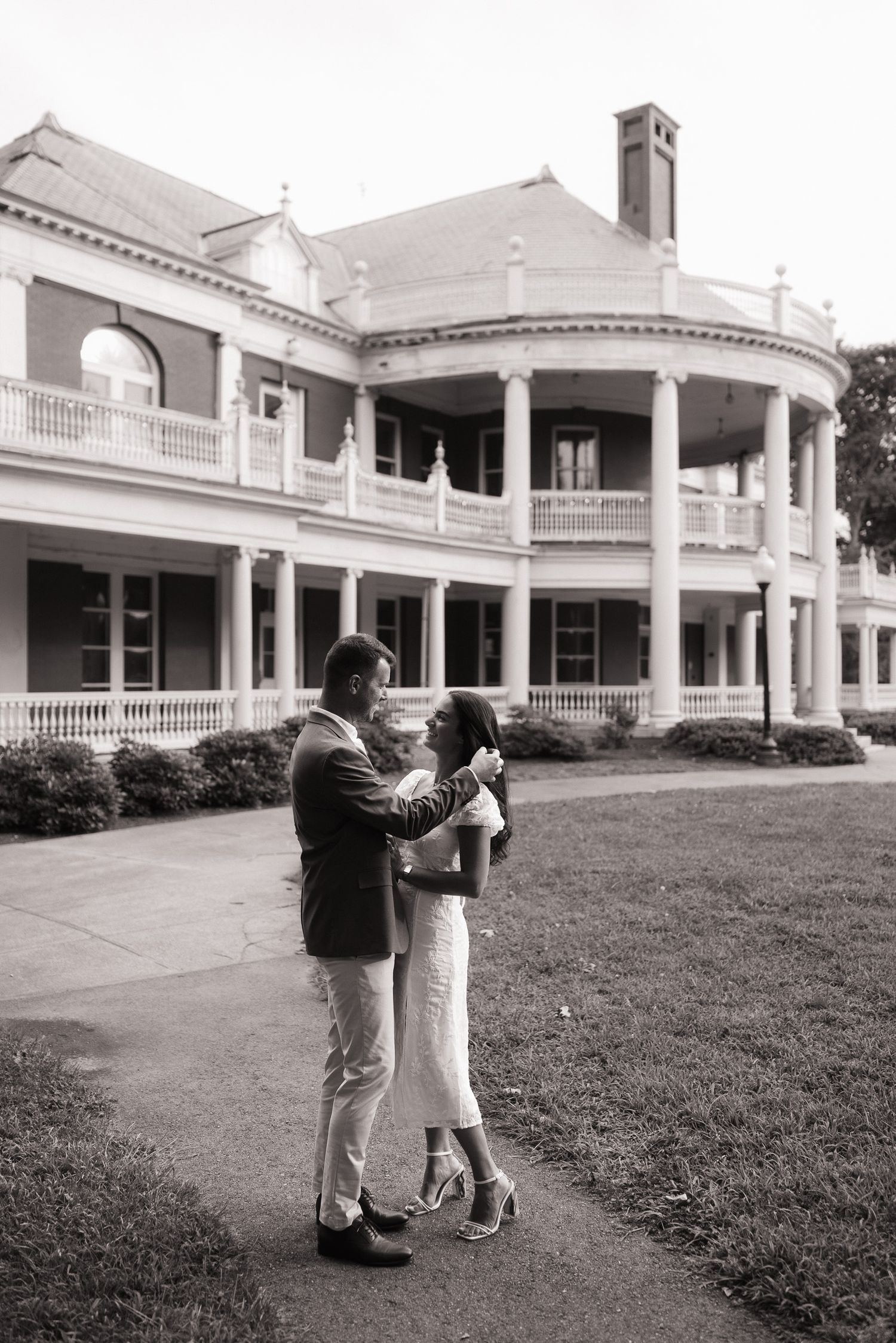 Black and white photo of a couple embracing outside a stately mansion with white columns.