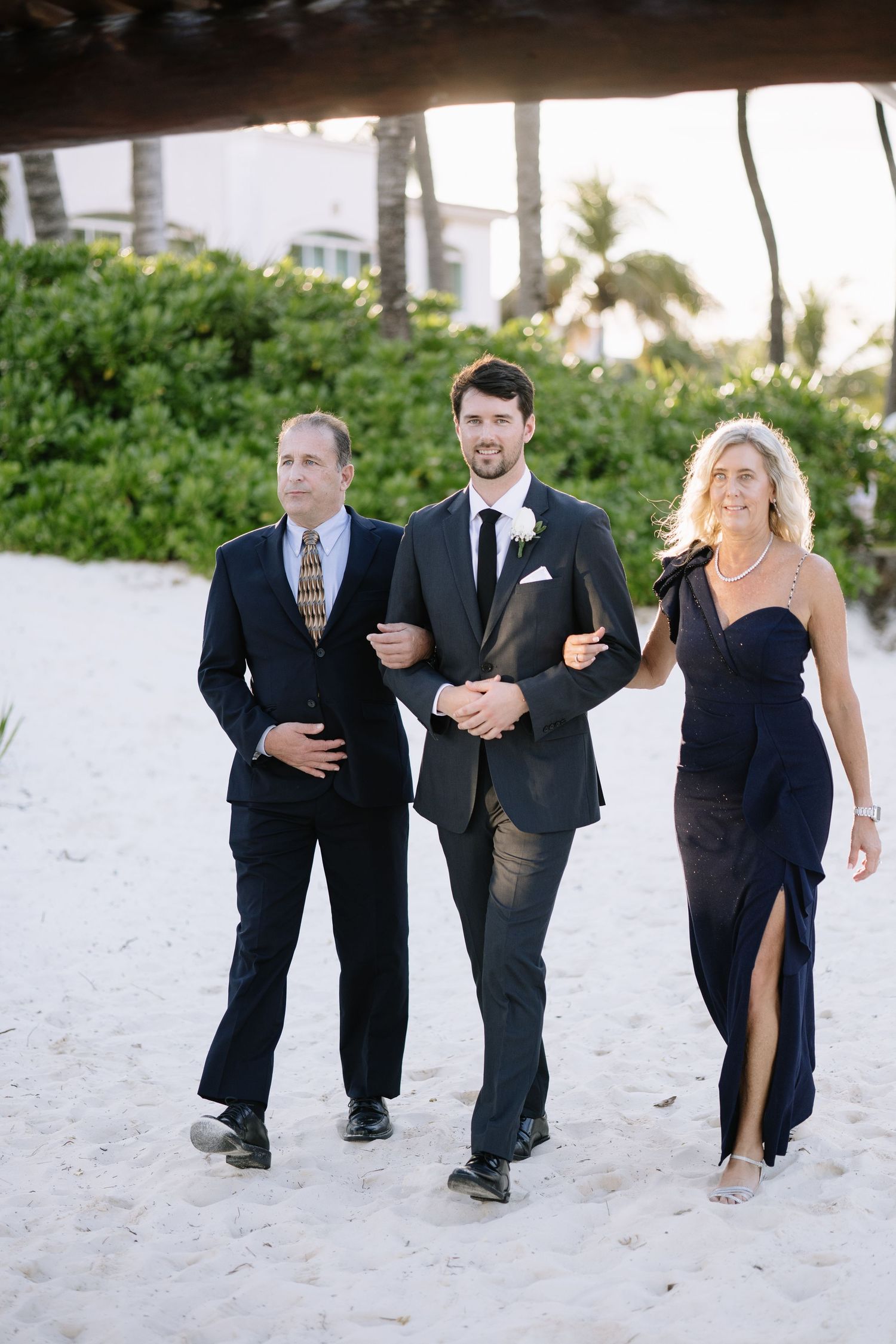 Wedding party walks along a sandy beach with palm trees and greenery in the background.