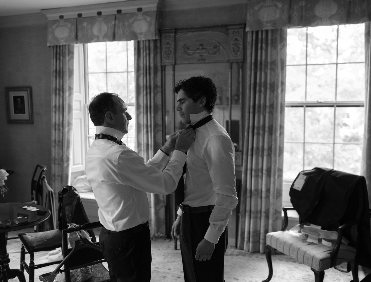 Black and white photo of two men in formal wear, one helping the other adjust his bow tie before a wedding.