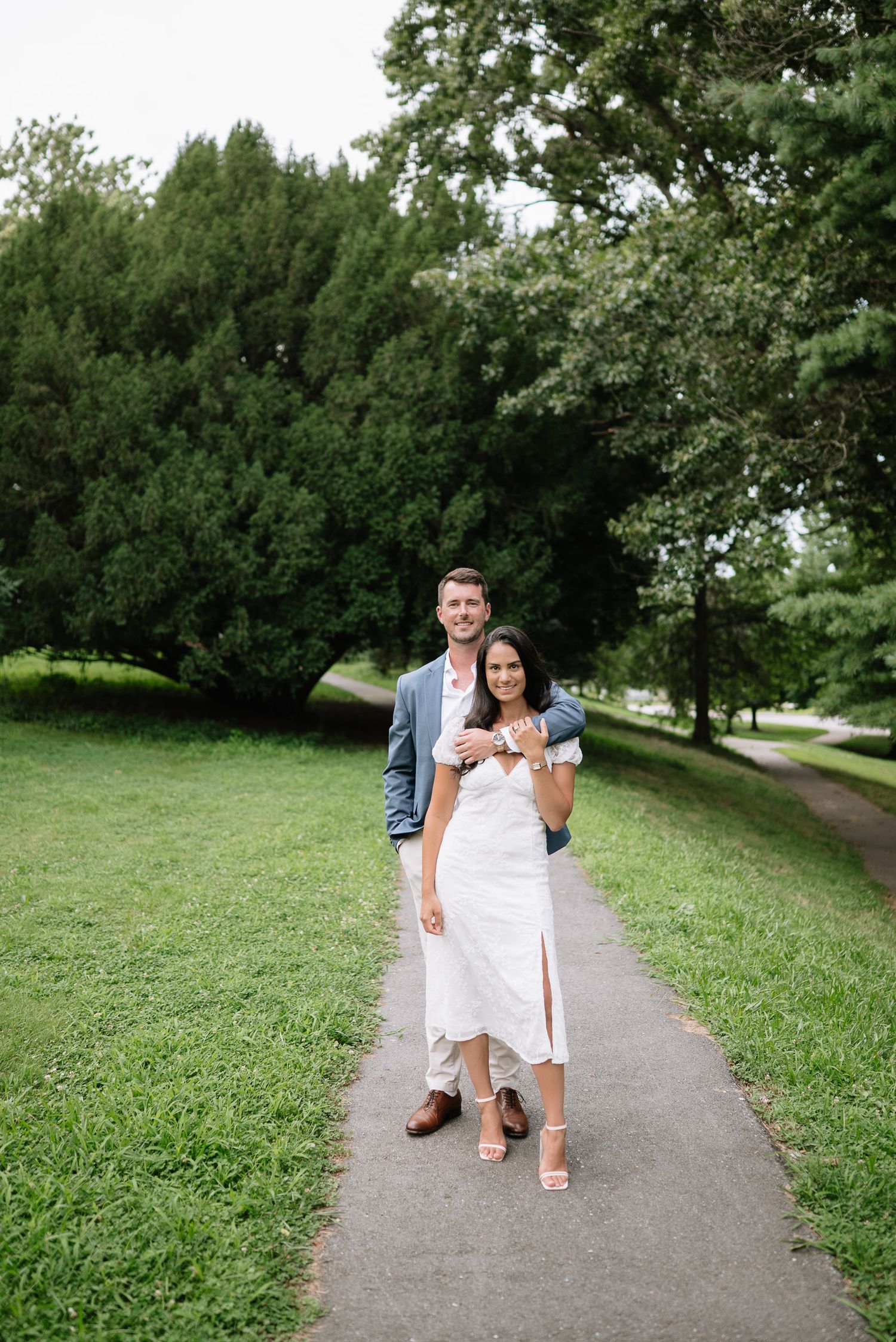 A couple strolls together on a winding park pathway surrounded by green grass and trees.