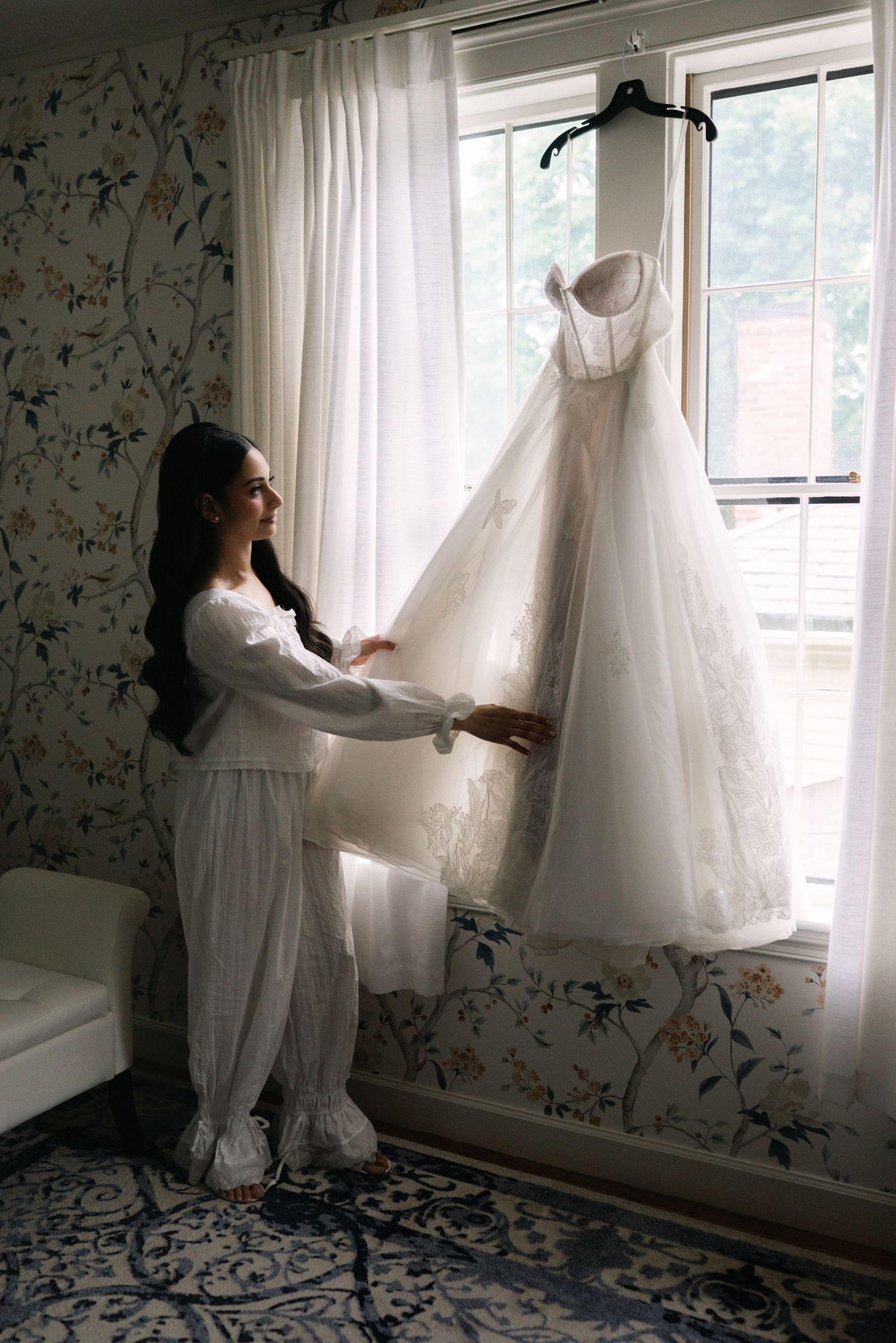 Bride in white pajamas admiring her wedding gown hanging by a sunlit window in a floral wallpapered room.