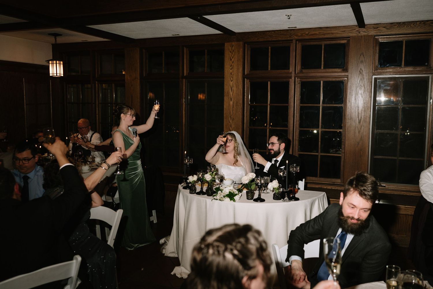 Wedding reception toast with bride and groom at sweetheart table, bridesmaid raising glass in elegant wood-paneled venue.