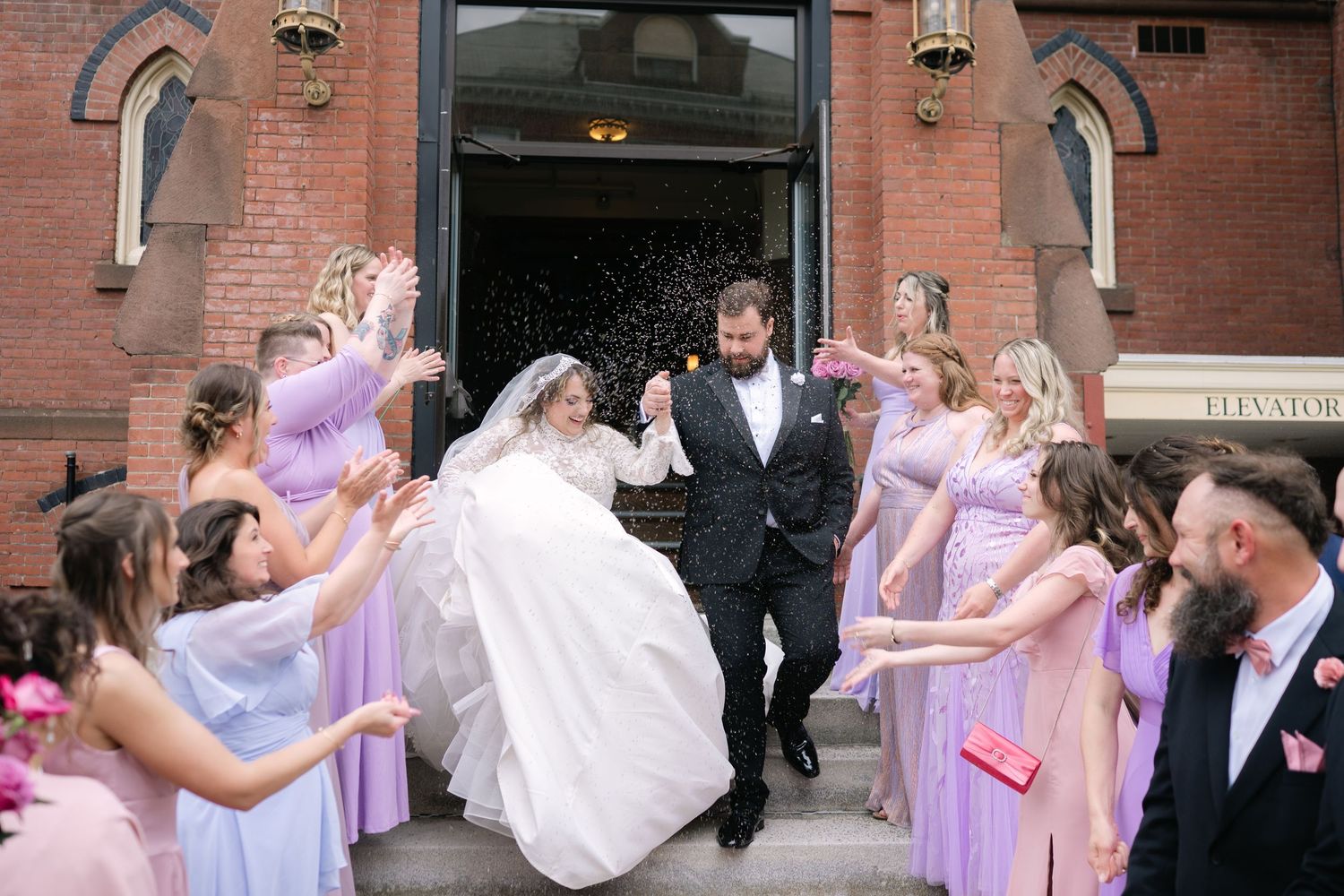 Wedding party celebrates with bubbles as newlyweds exit brick church in lavender and white attire.