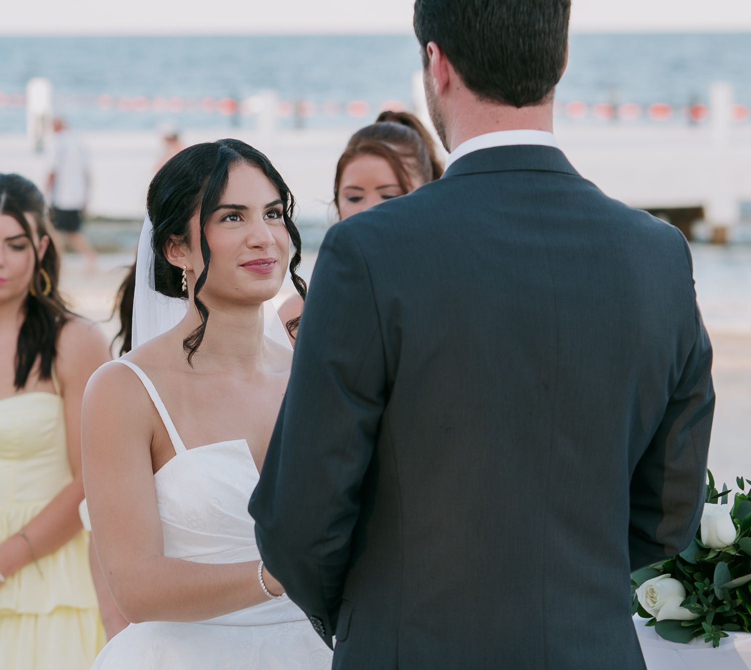 A bride in a white wedding dress gazes at her groom during an outdoor beach ceremony.