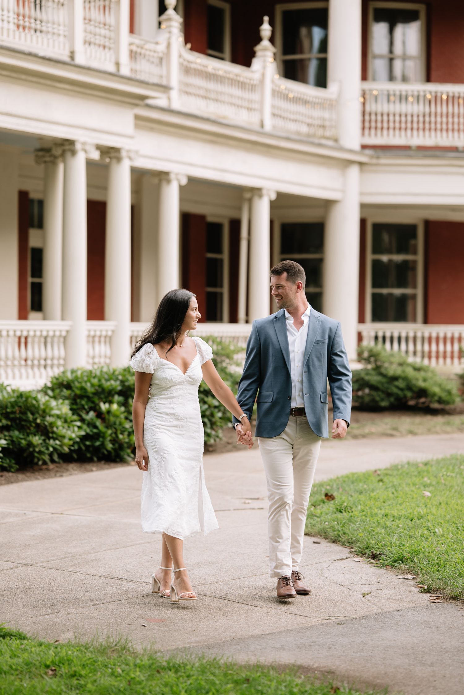 A couple in summer formal wear walks playfully along a historic building's walkway.