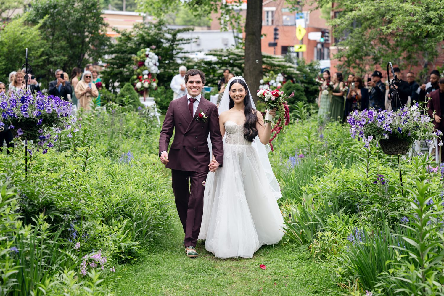 Bride and groom walk through lush garden aisle surrounded by purple wildflowers and greenery on their wedding day.