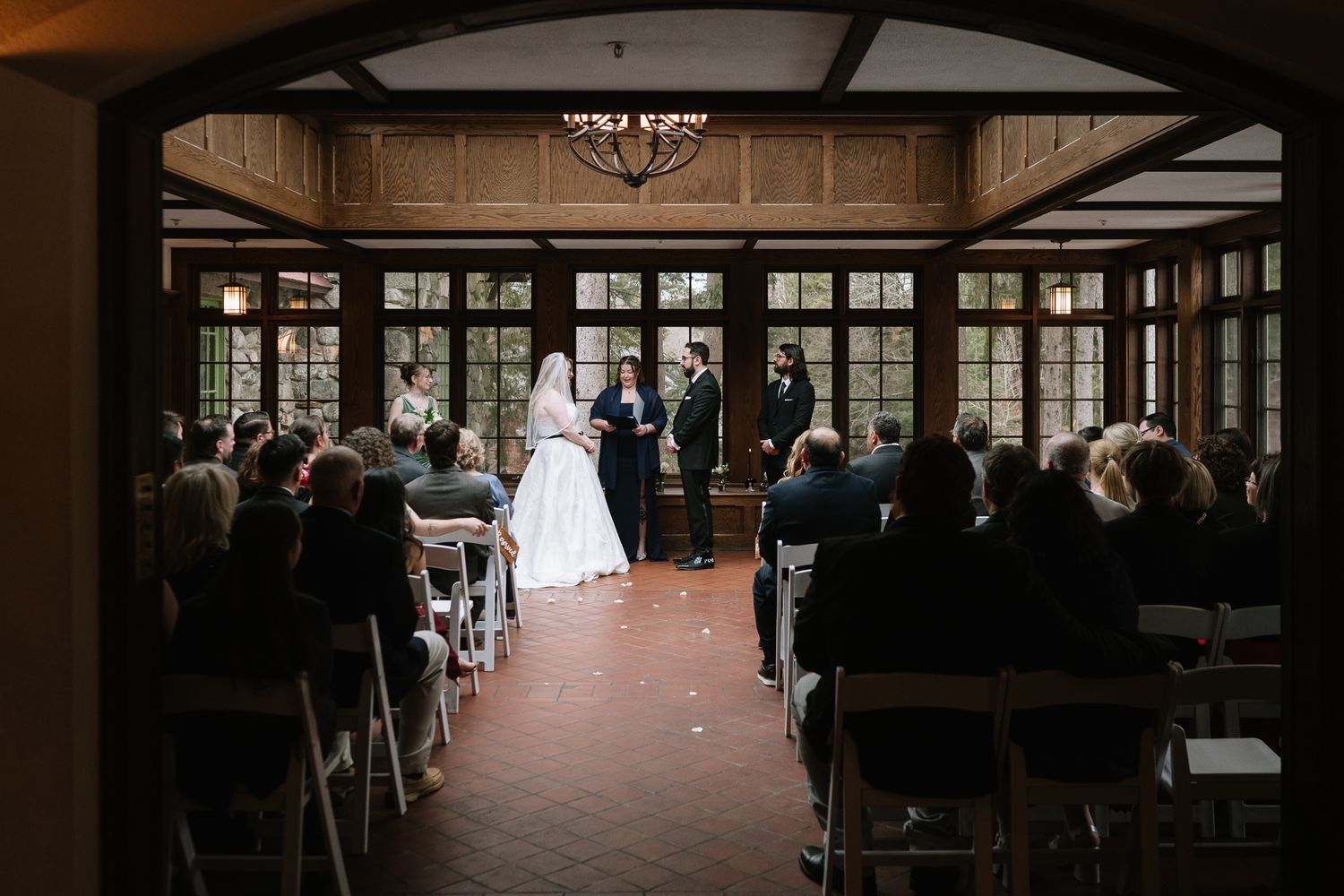 Elegant indoor wedding ceremony in a rustic wood-paneled venue with guests seated in white chairs facing the altar.