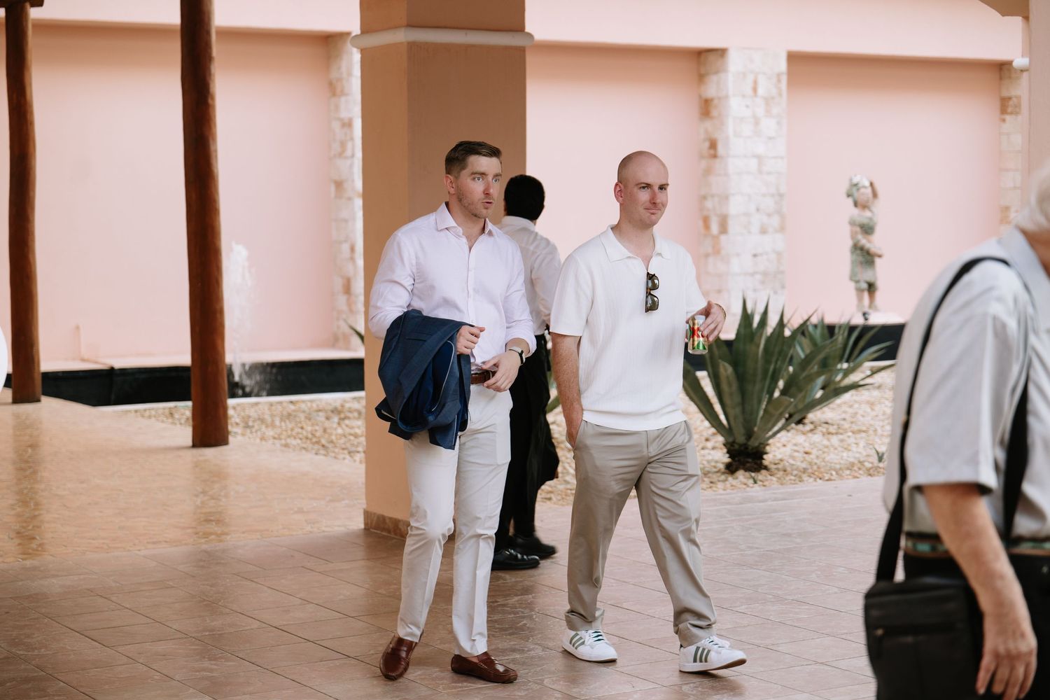 Wedding guests walk through a covered walkway with pink walls and tropical landscaping.