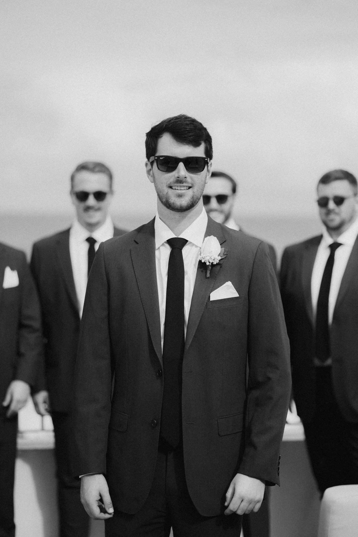 Groomsmen in dark suits and sunglasses pose for a black and white wedding photo.