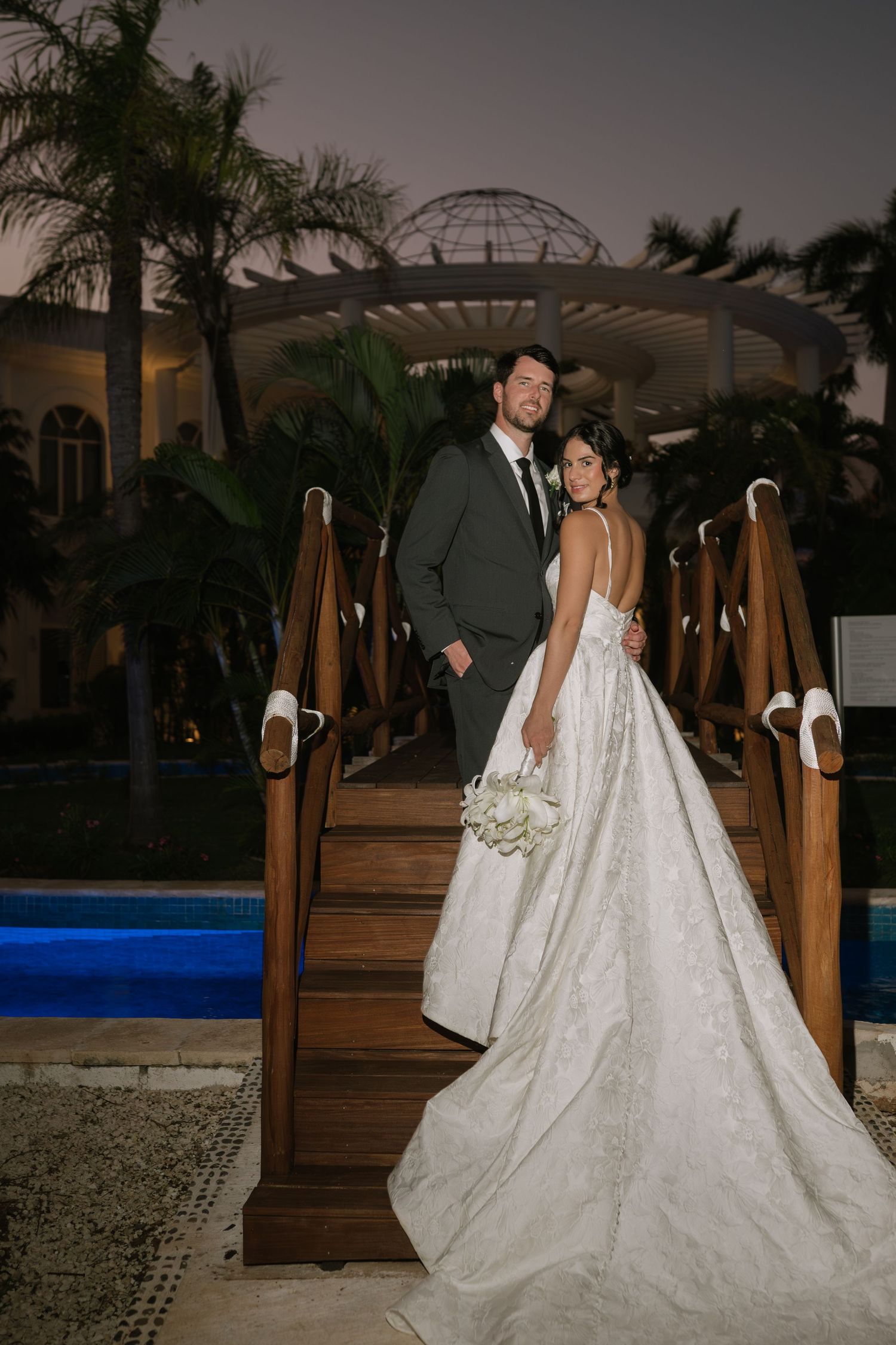 A romantic wedding couple poses together on a bench at dusk with decorative lighting and palm trees in the background.