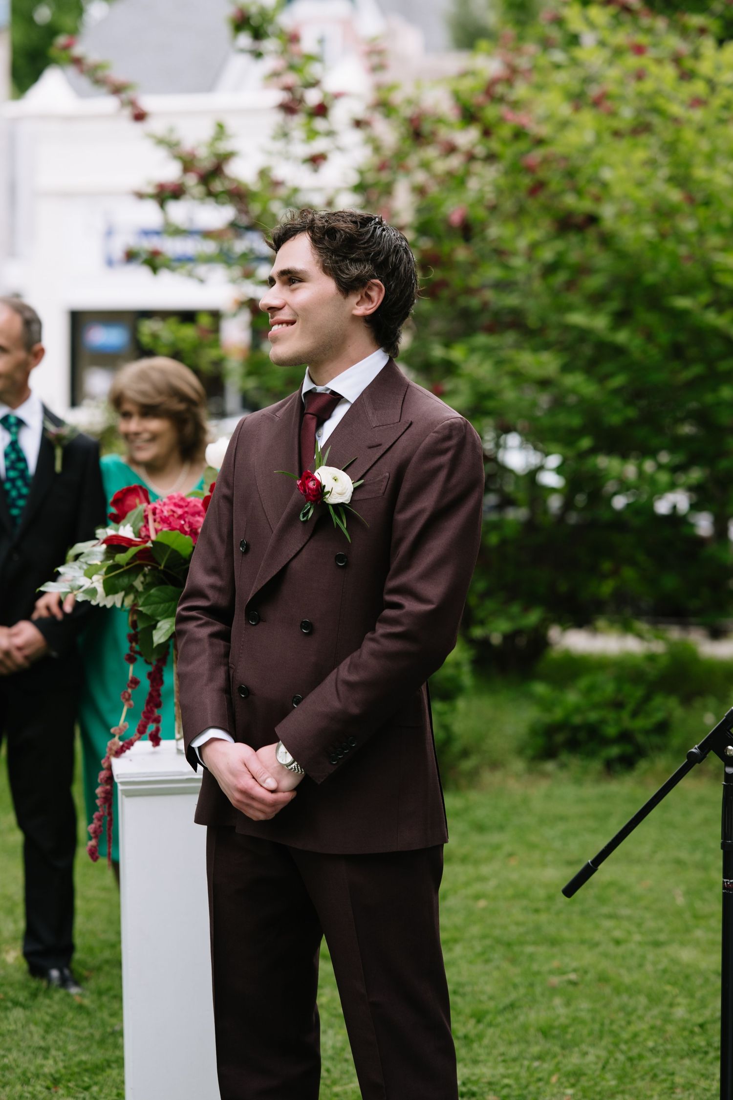 Groom in brown double-breasted suit with white boutonniere smiling at outdoor wedding ceremony with green garden backdrop.