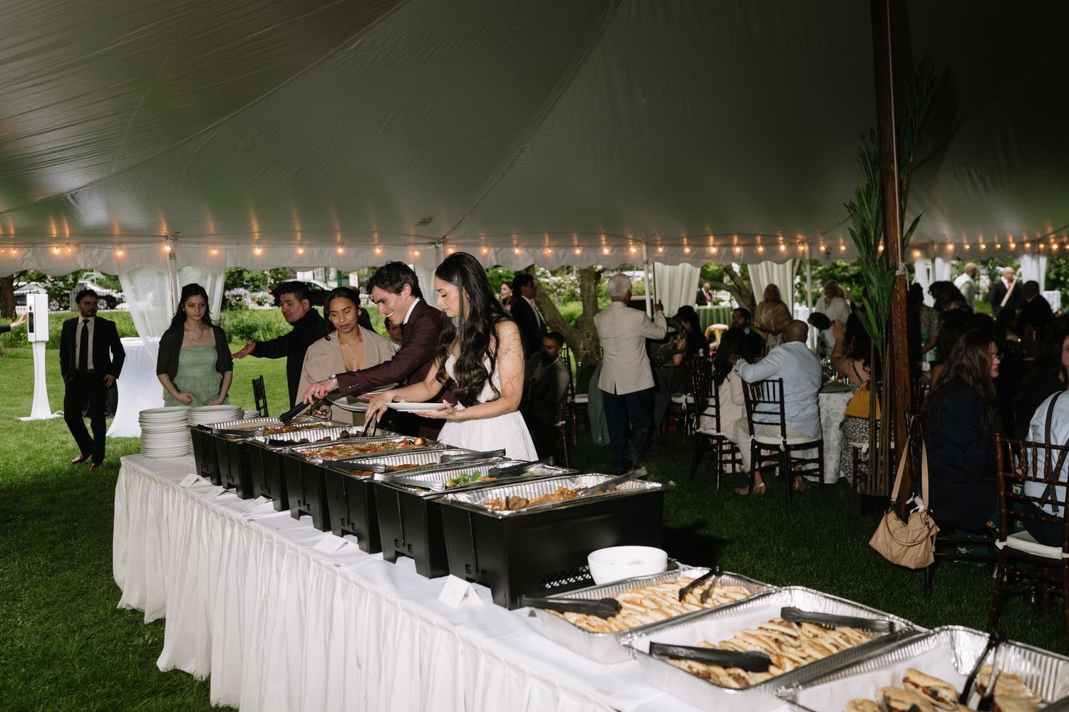 Guests serving themselves at an elegant outdoor wedding buffet under a white tent with string lights.