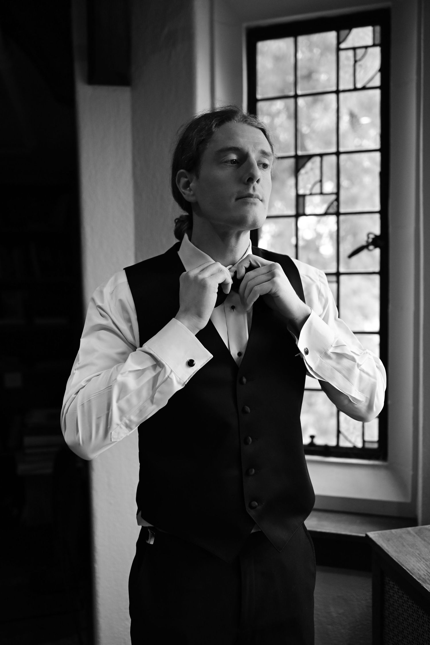 A person adjusts their bow tie while wearing formal attire near a window in a black and white photo.