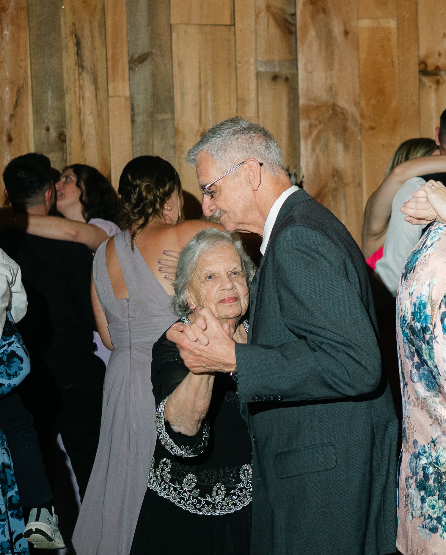 Wedding guests dancing together at an indoor reception with wooden walls and floral dresses.
