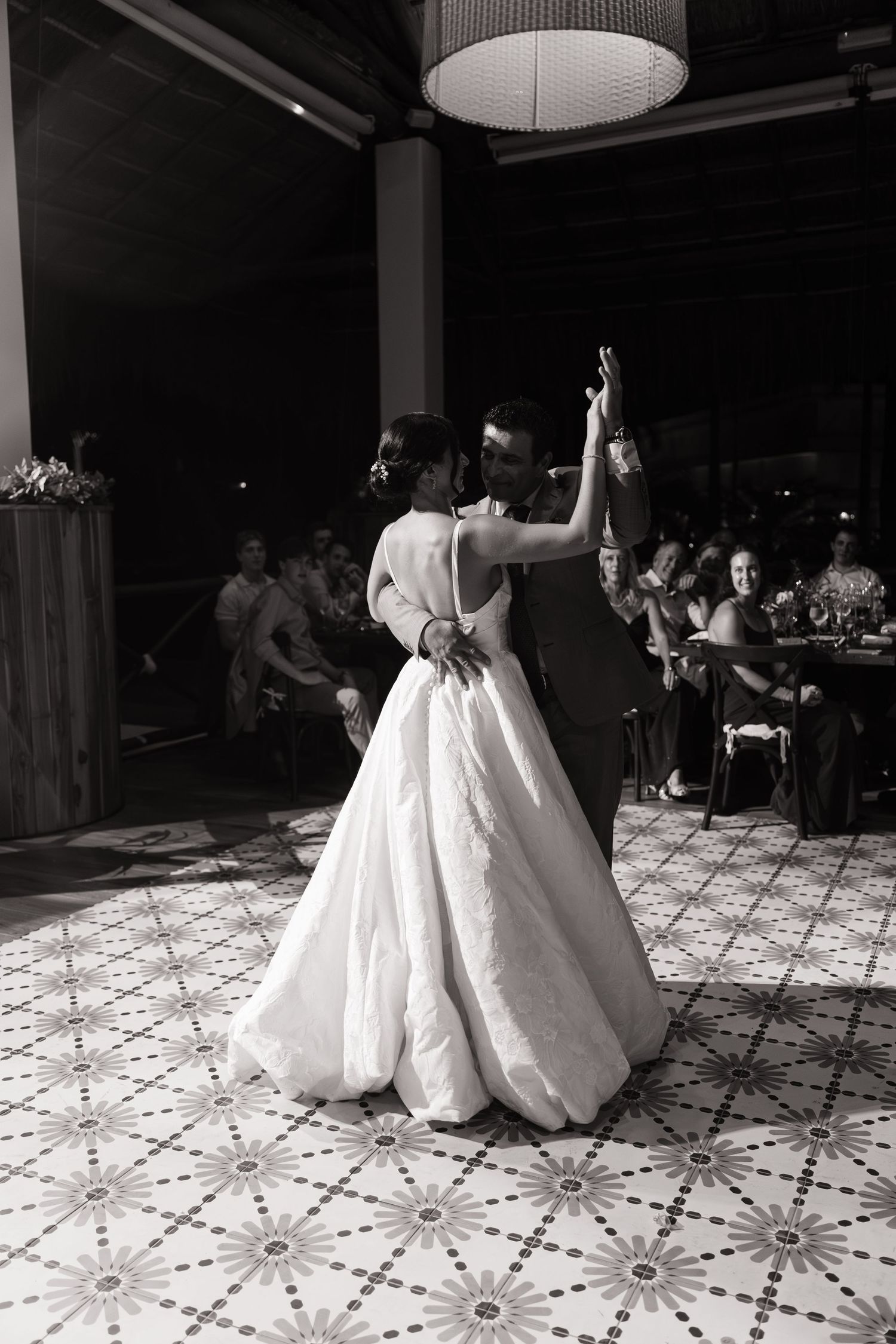 A couple shares their first dance at a wedding reception on a patterned dance floor under dim lighting.