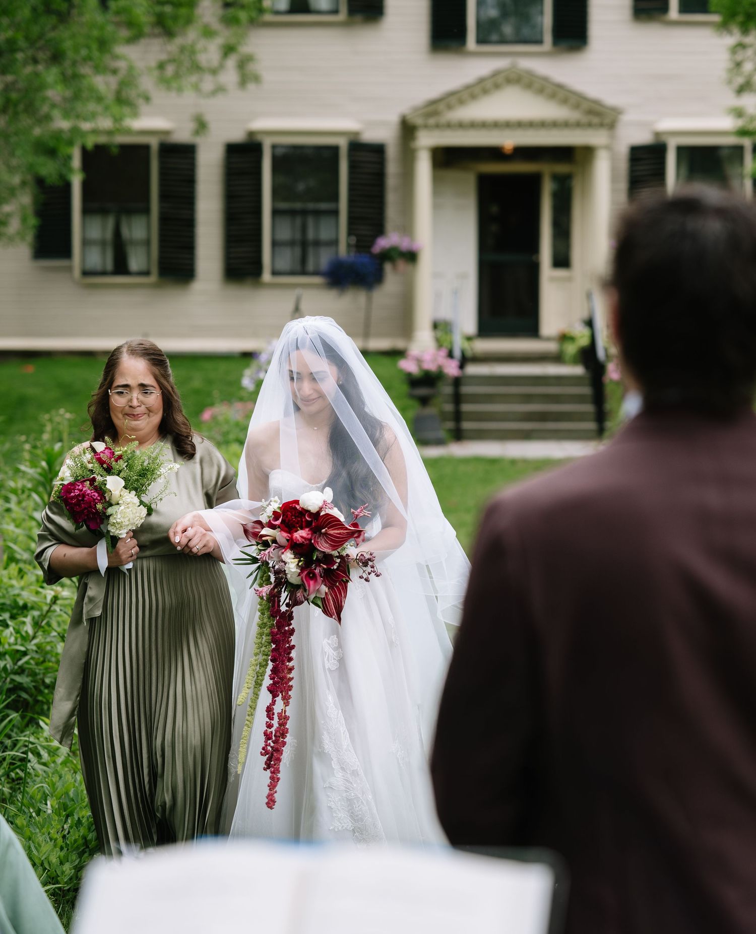 Bride in white gown and veil walks with bridesmaid in green dress toward altar at outdoor wedding ceremony.