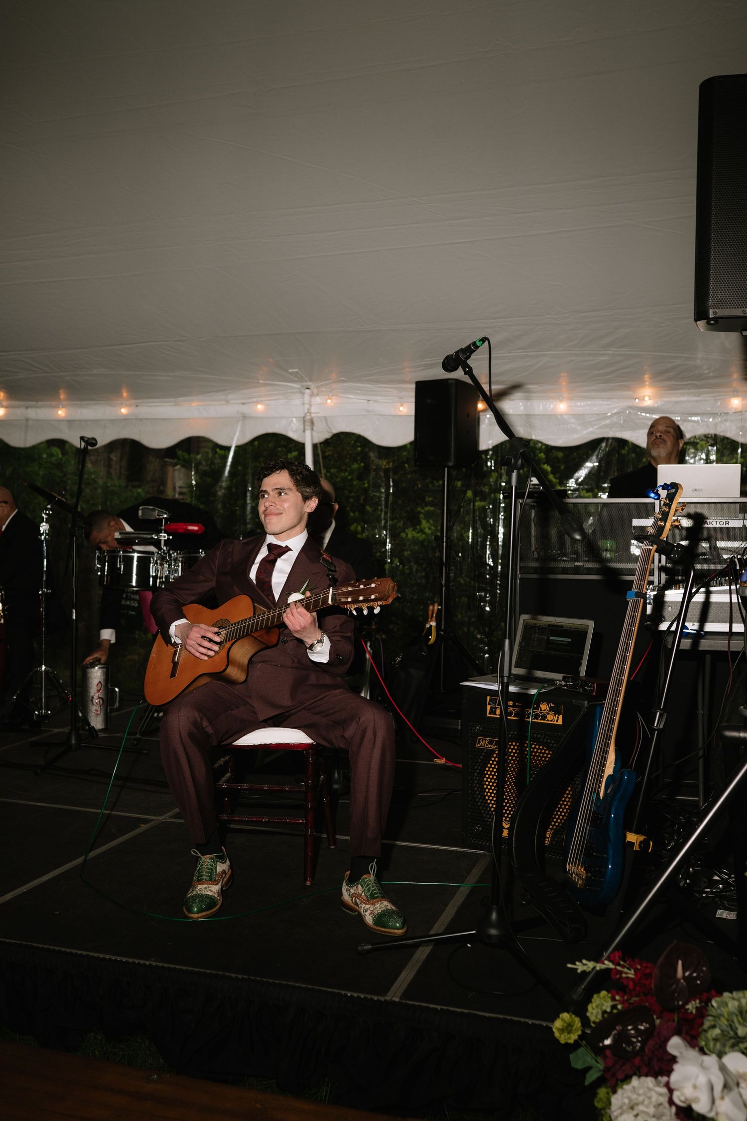 A musician in a brown suit plays acoustic guitar on stage at an elegant tented event.