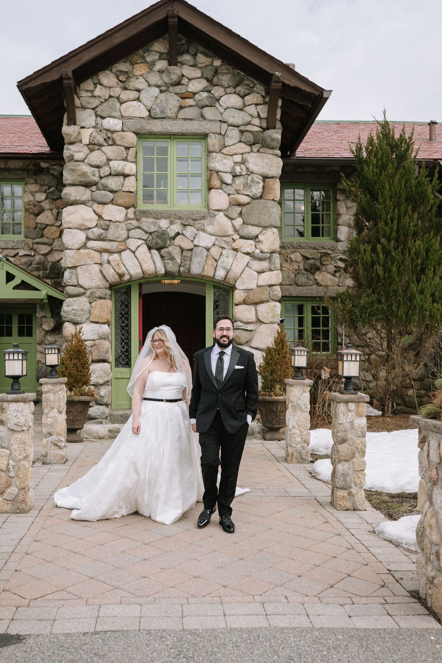 Bride and groom holding hands in front of a rustic stone building venue on their winter wedding day.