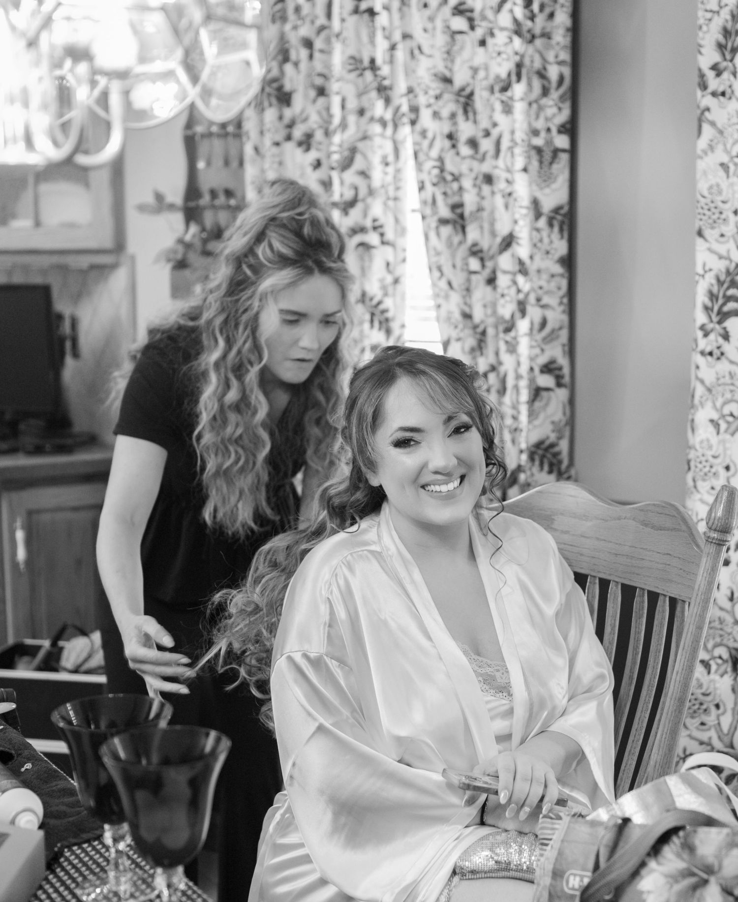 Black and white photo showing a hairstylist working on a client wearing a white silk robe during wedding preparations.