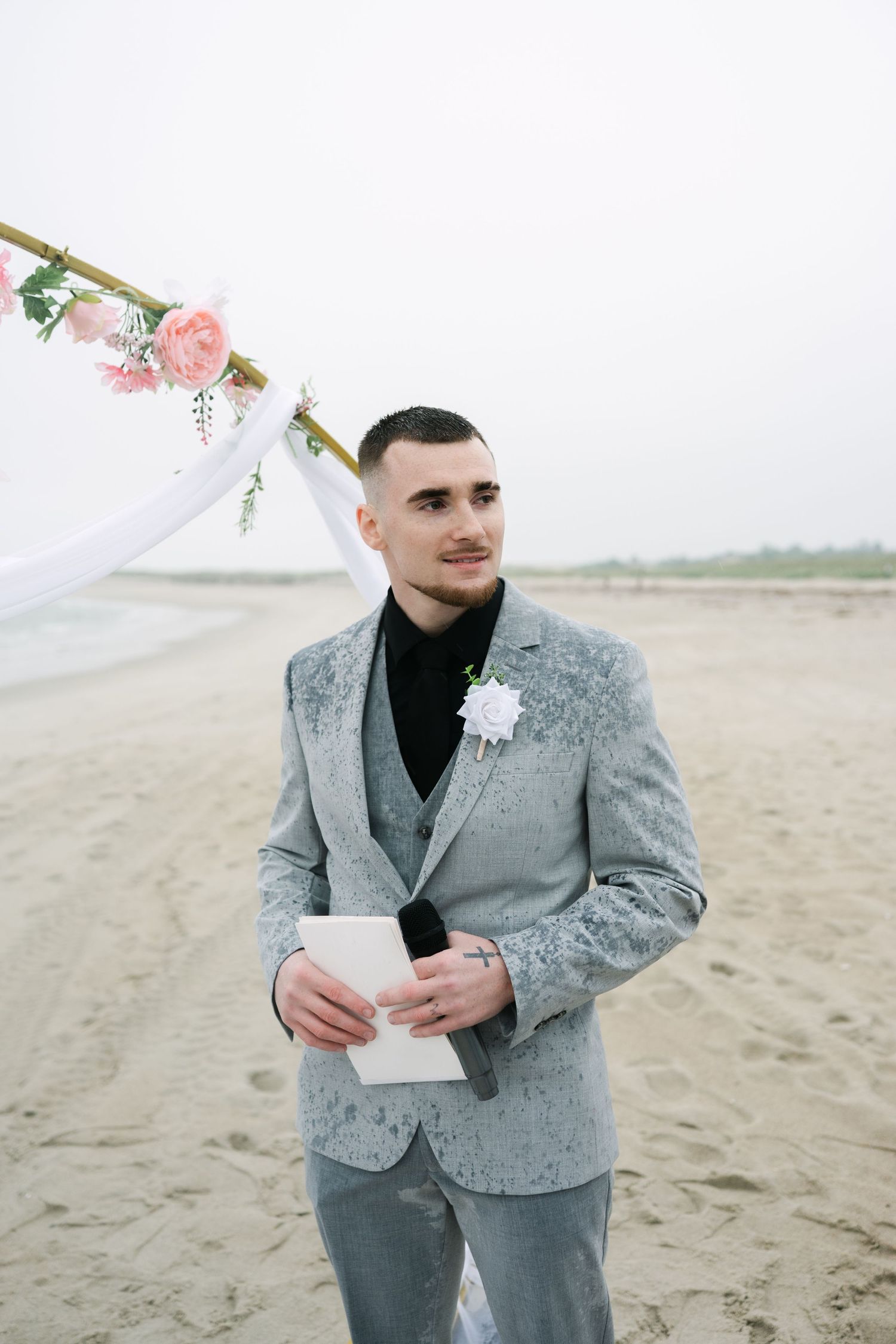 A person in a stylish gray suit jacket reads a letter while standing on a sandy beach.