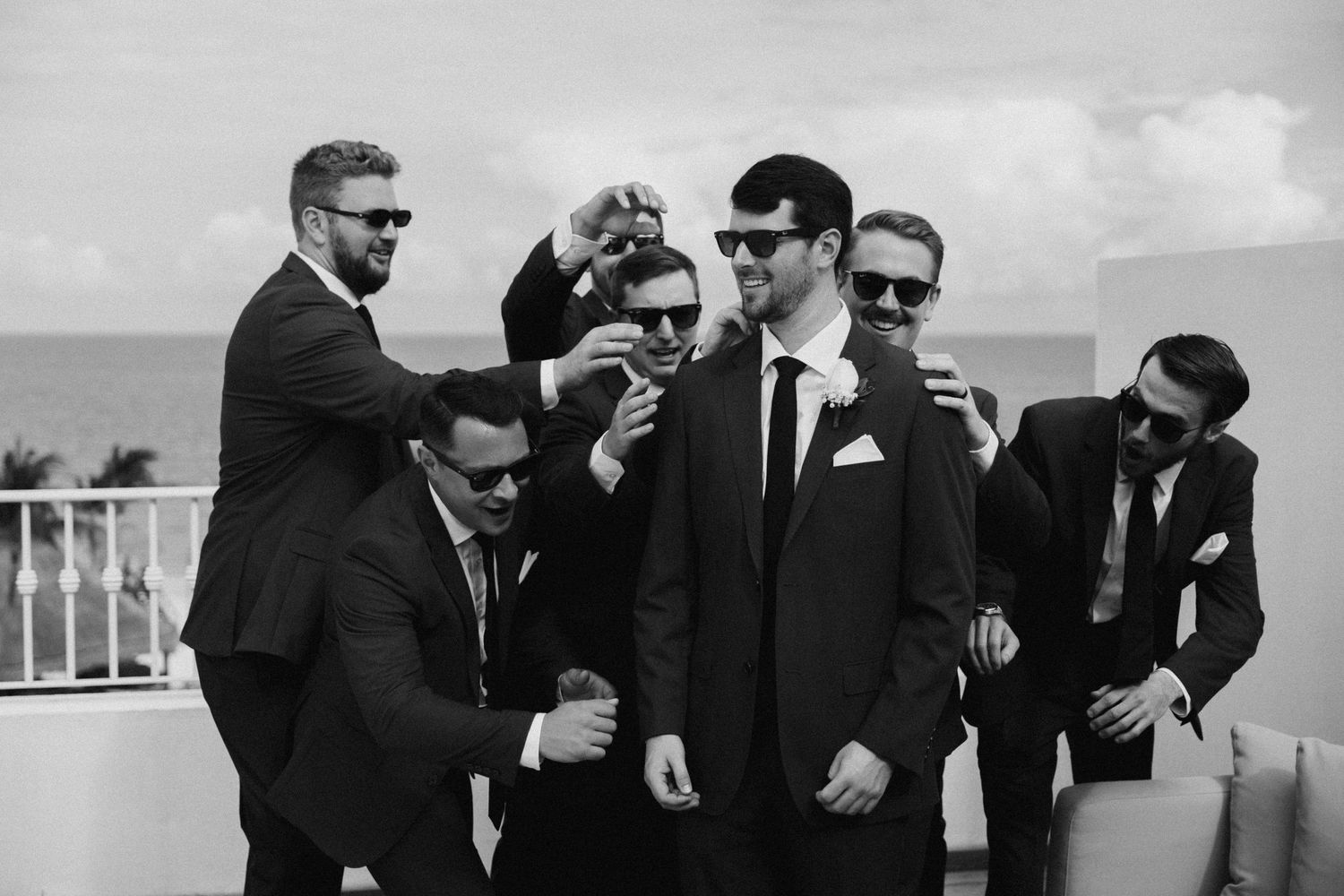 Groomsmen in black suits and sunglasses share laughs while posing together outdoors.