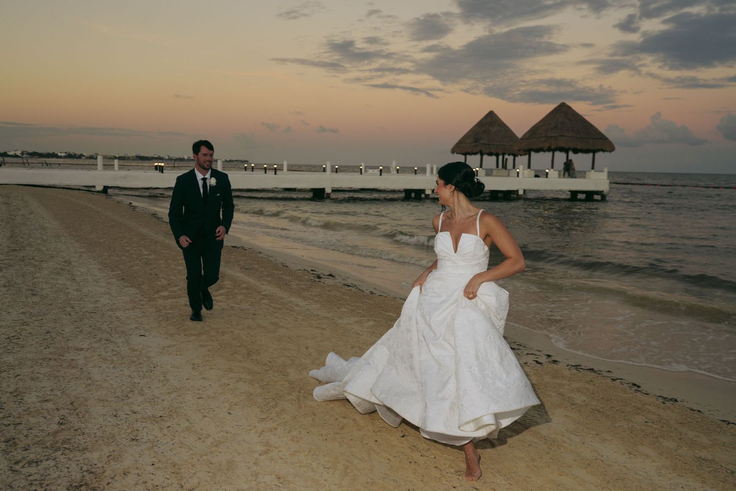 A couple in wedding attire walks along a tropical beach at sunset with overwater bungalows in the background.