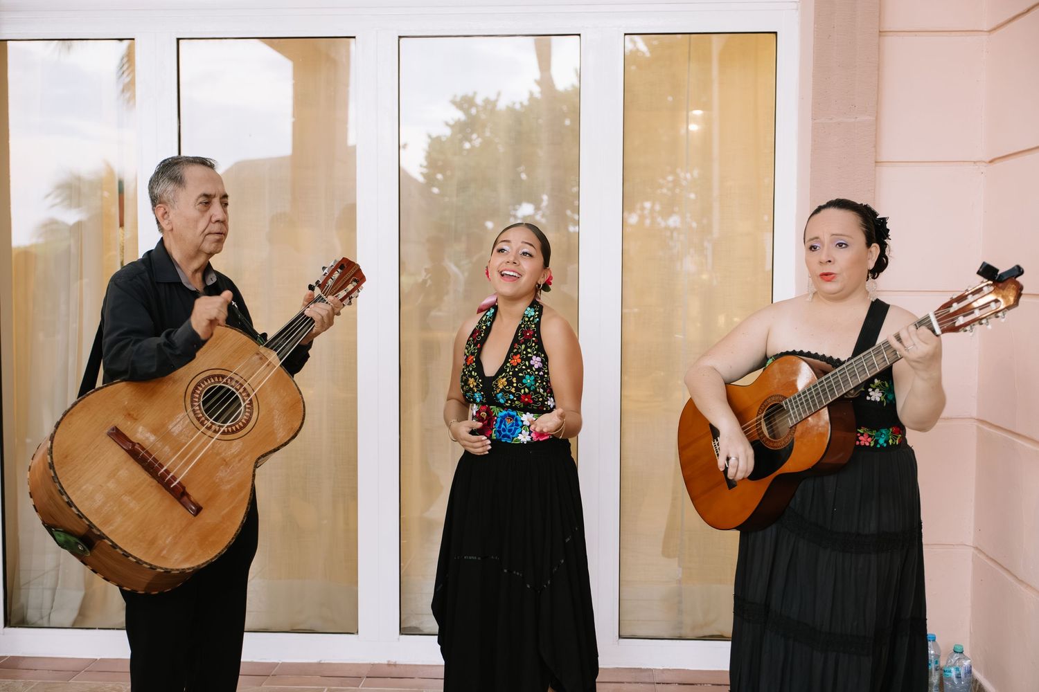 Three musicians perform together with acoustic guitars and vocals in front of large windows.