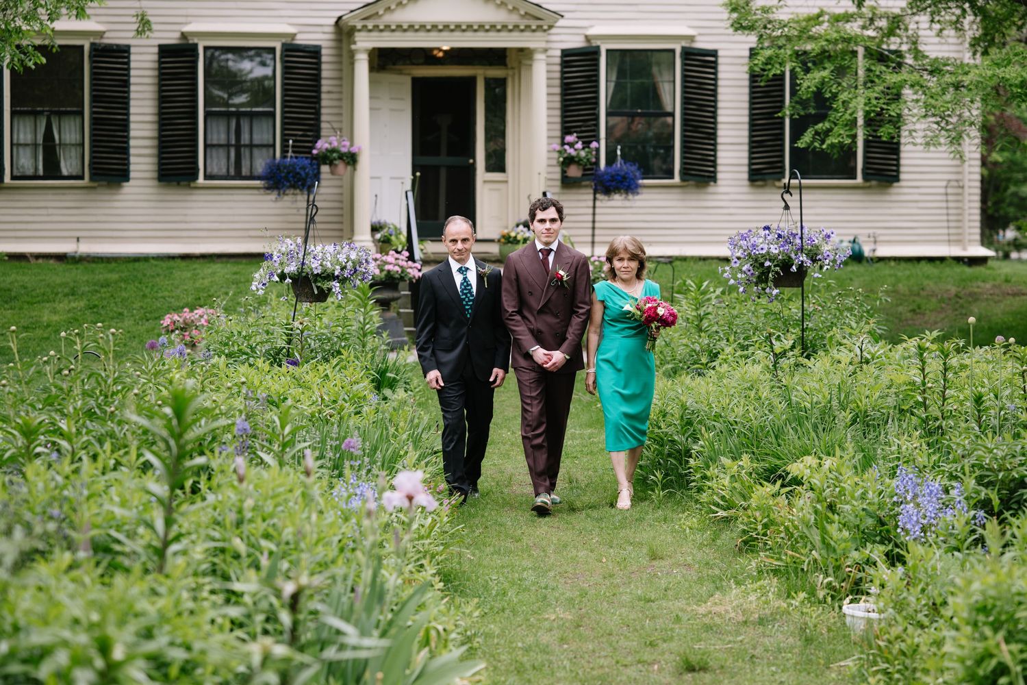 Groom walks with parents through lush garden path toward white house during outdoor wedding ceremony.