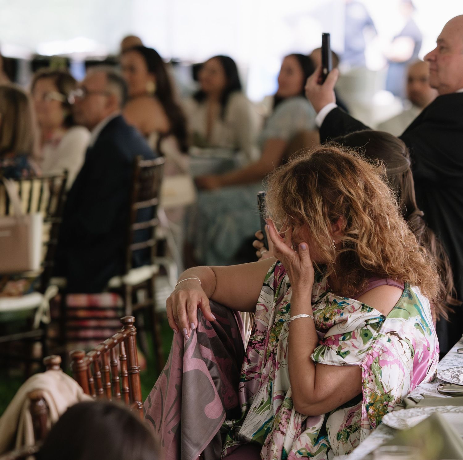 Guests seated at an elegant outdoor event under a white tent, with a woman in a floral dress in the foreground.