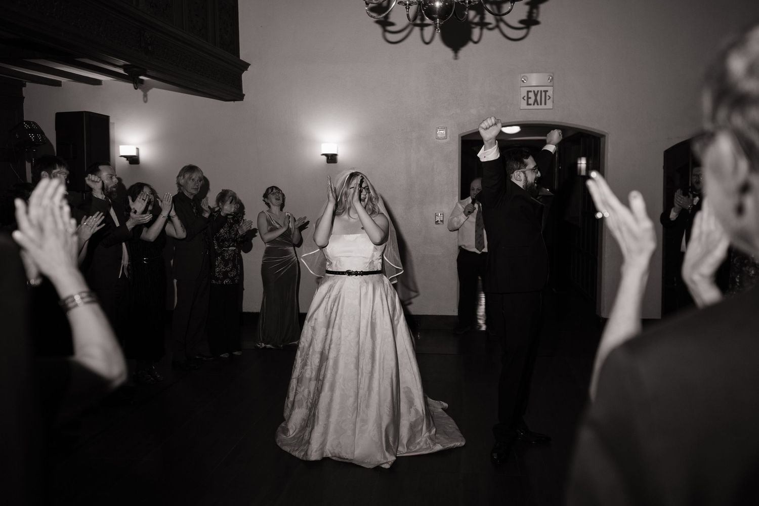 Black and white photo of bride and groom entering wedding reception as guests clap and celebrate.
