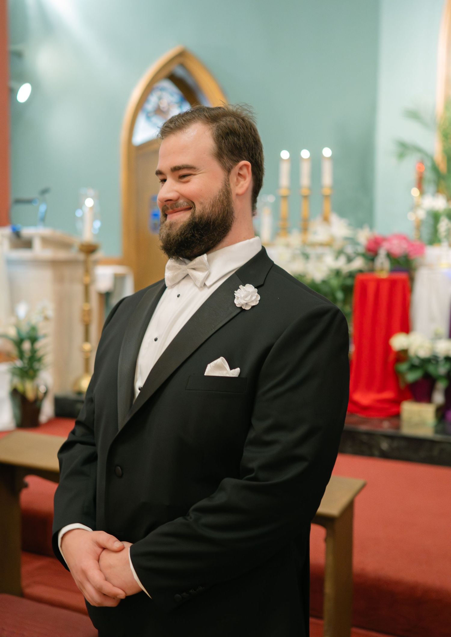 Groomsmen in black tuxedos and bow ties stand in a church sanctuary with candles.