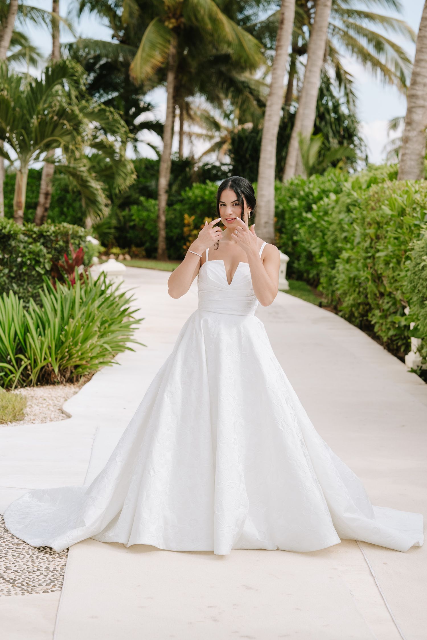 Bride in elegant white ball gown poses on tropical path with palm trees.