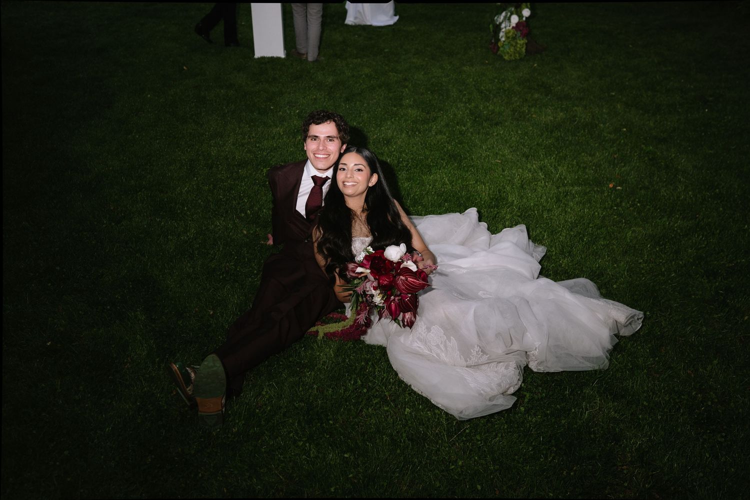 Bride and groom sitting on green grass, smiling, bride in white gown holding red bouquet, groom in dark burgundy suit.